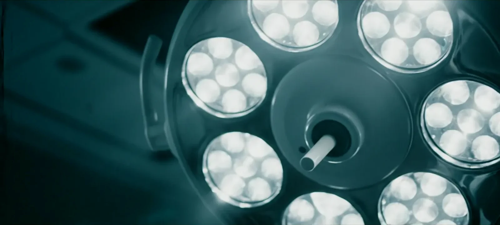 A surgical theatre light seen from below, its lamp heads glowing in cool teal.