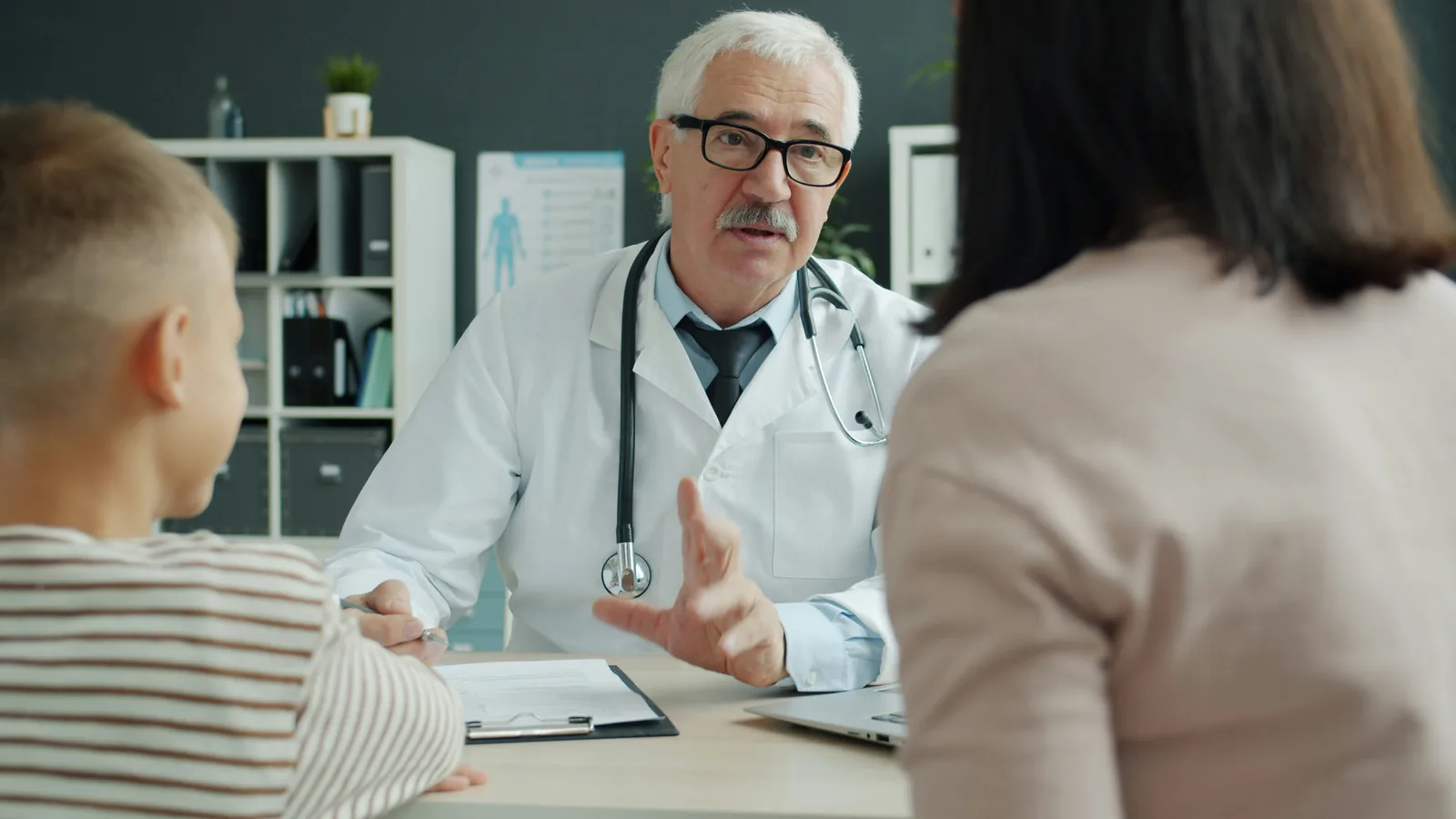 An older doctor in a white coat with a stethoscope around his neck sits at a consulting room desk, one hand raised mid-explanation, talking with a woman and a child seen from behind, a clipboard and a laptop on the desk and shelves of binders behind him