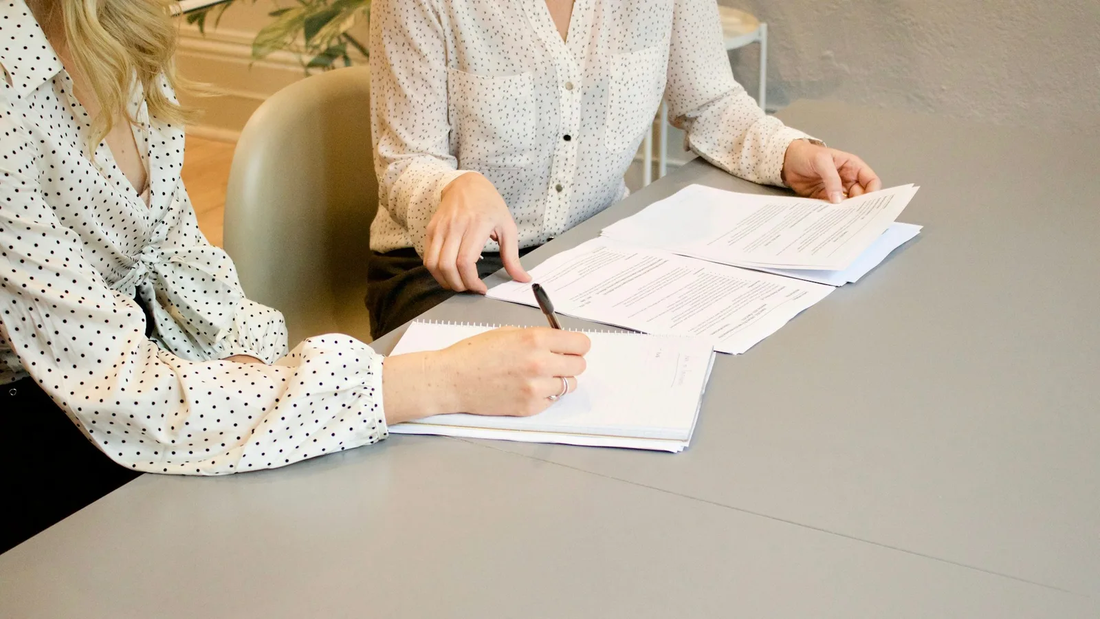 Two people seated at a grey table working through spread-out printed documents, one holding a pen over an open notebook