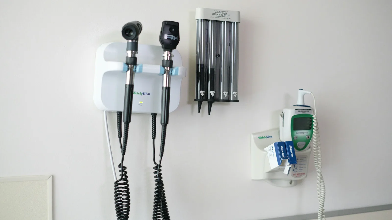 A wall-mounted otoscope and ophthalmoscope diagnostic set, specula dispenser and ear thermometer on the white wall of a GP examination room