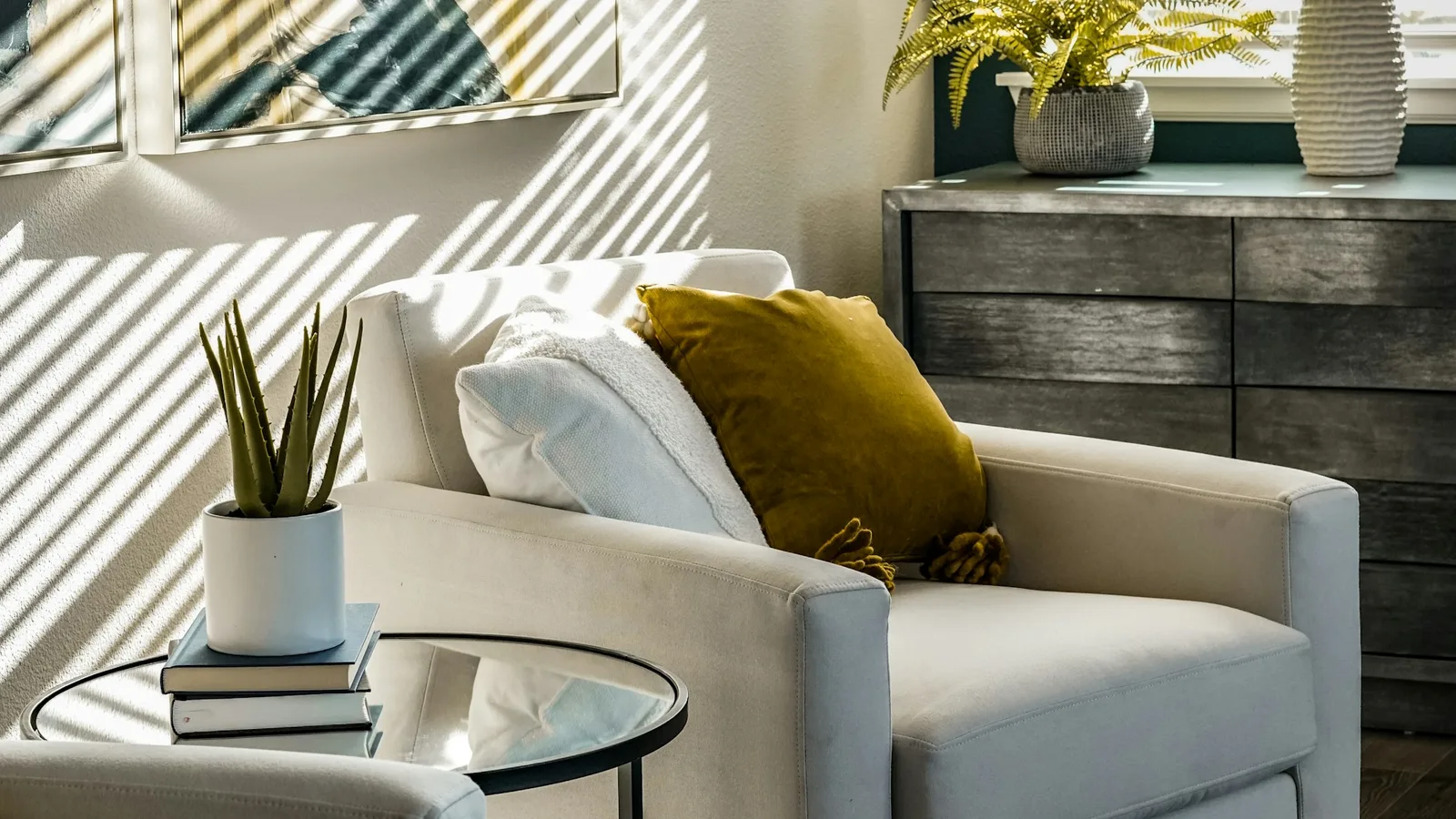Two pale armchairs and a glass side table in a calm sunlit room, blind shadows falling across the wall