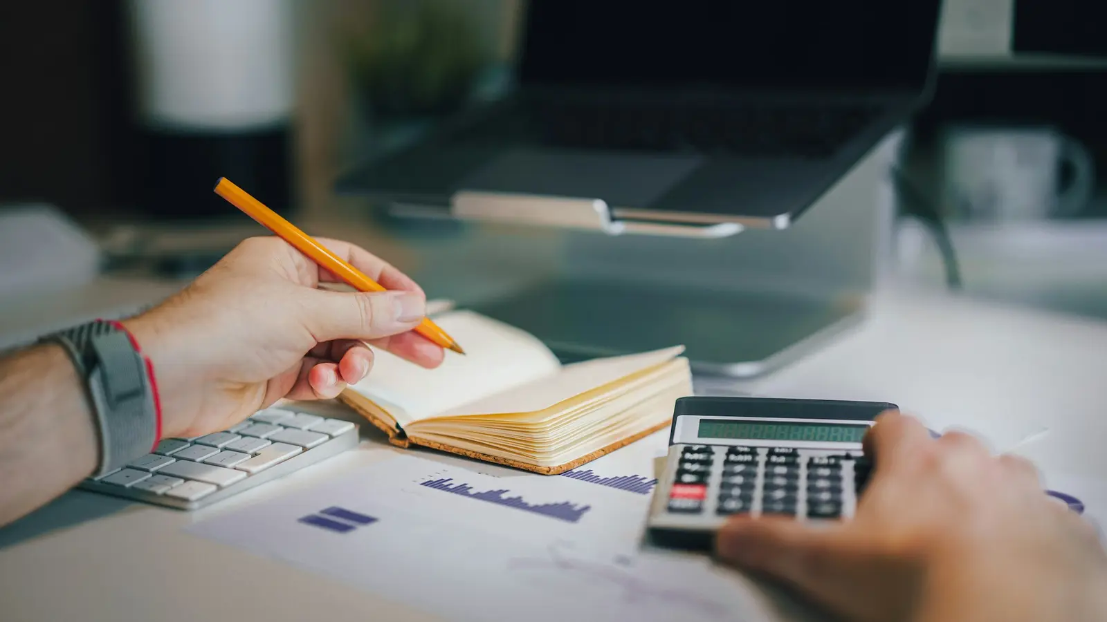 A person's hands work at a calculator beside an open laptop and notebook on a pale desk in soft natural light