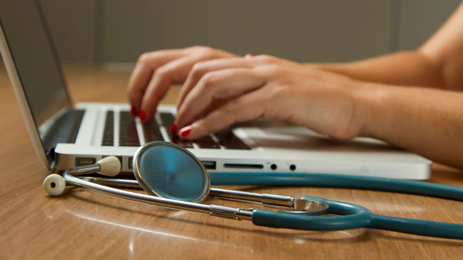 A clinician's hands typing on a laptop keyboard beside a teal stethoscope on a wooden desk