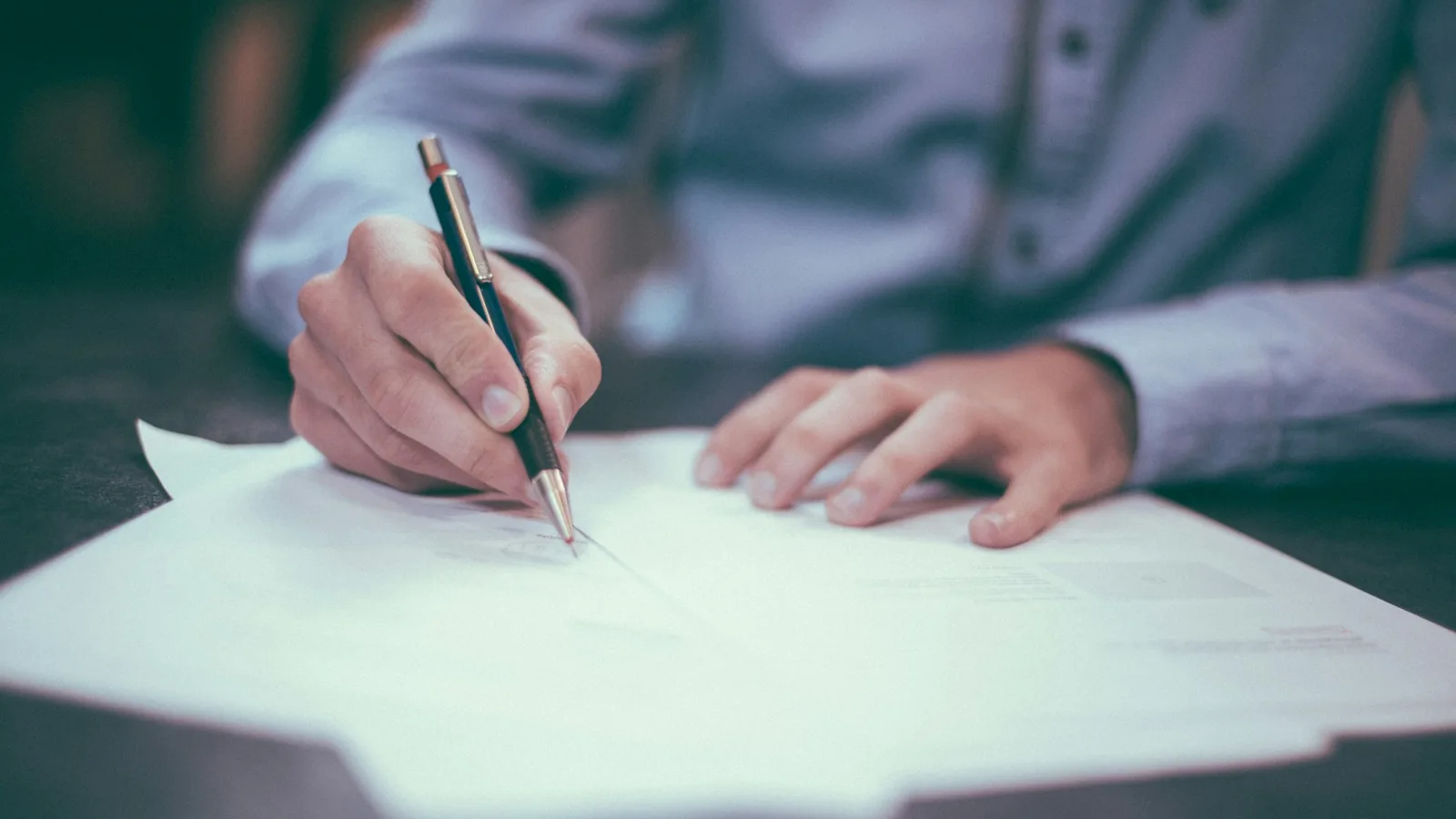 Close-up of a person's hands in a blue shirt signing paperwork with a pen, documents spread across a dark desk