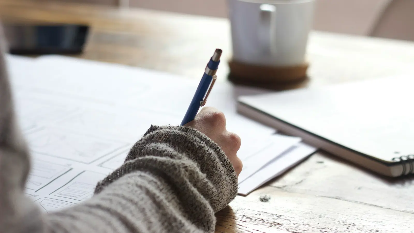 A hand works through printed forms with a pen, coffee cup alongside