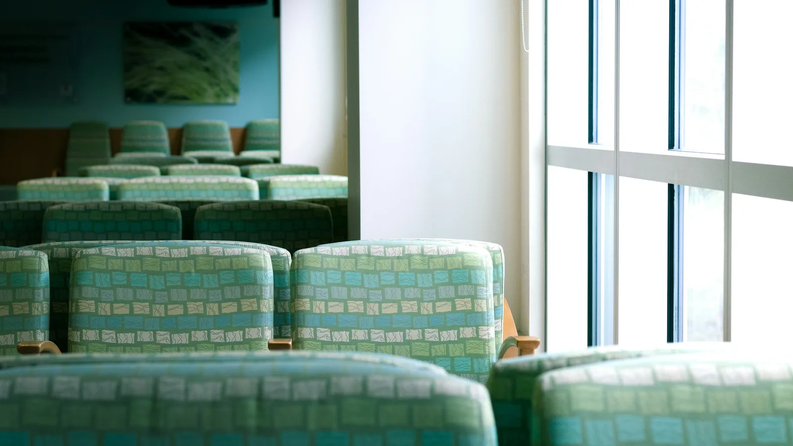 Rows of empty teal upholstered chairs in a quiet clinic waiting room beside a bright window