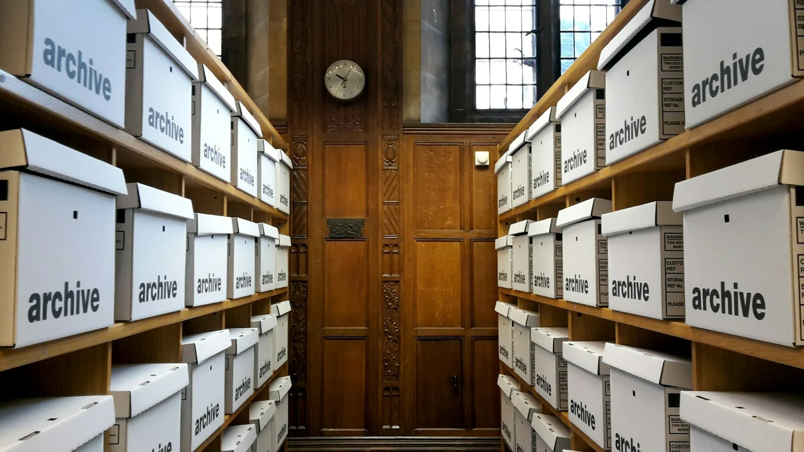 Rows of labelled archive boxes on wooden shelves flanking a wood-panelled wall with a clock and leaded windows