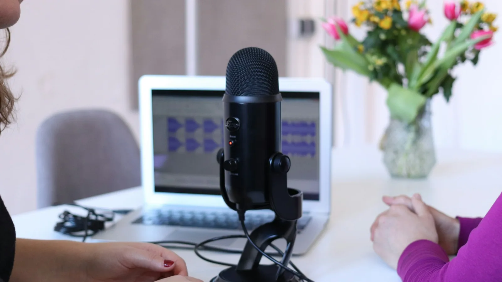 Two people sit across a table with a microphone between them and a recording waveform on a laptop screen