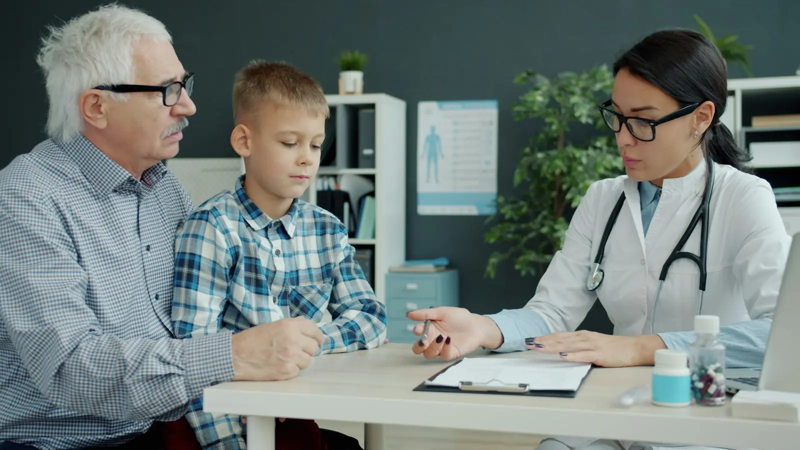 A clinician talks with a patient and a family member around a consulting room desk beside a bright window