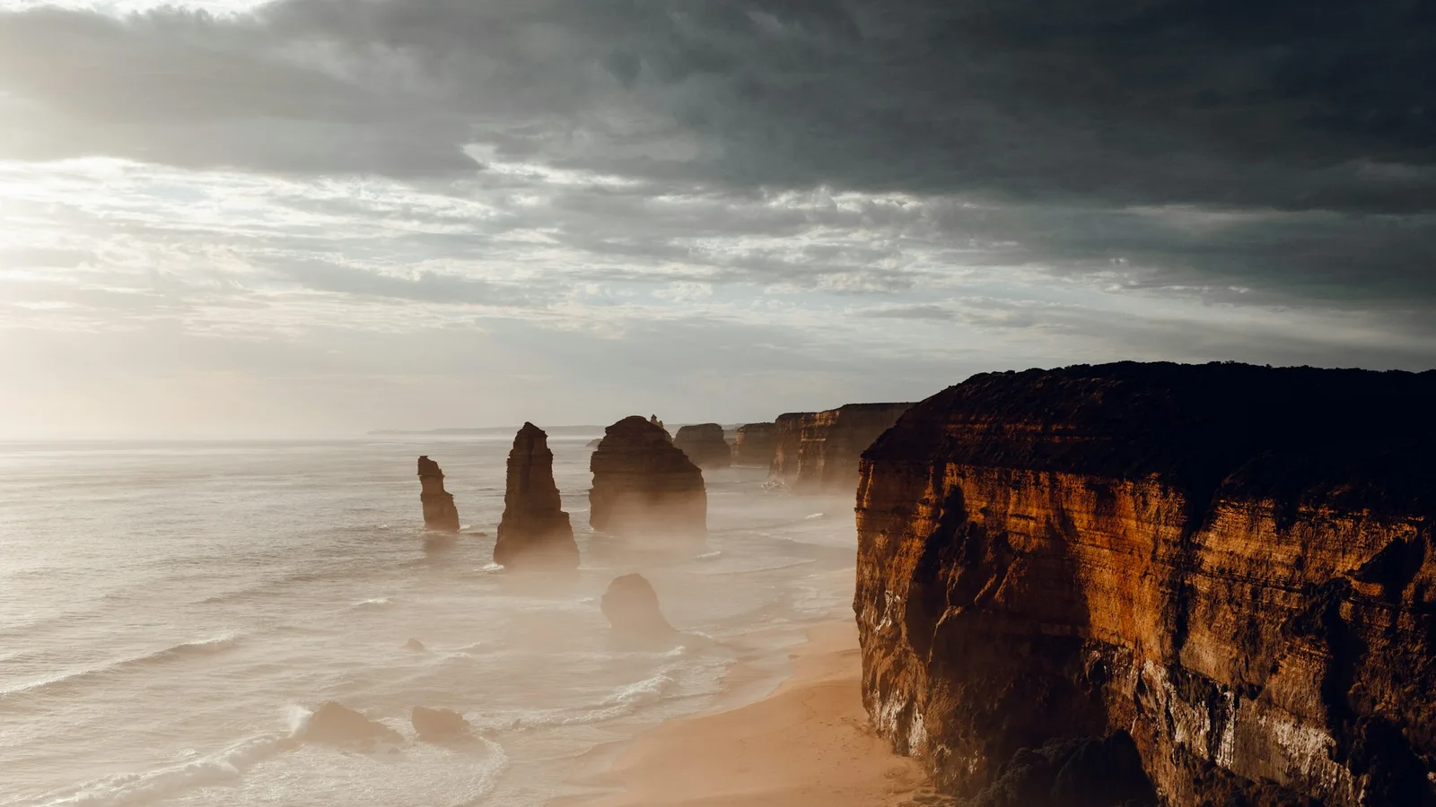 Limestone sea stacks of the Twelve Apostles rise from misty surf beside golden cliffs at dusk on Victoria's Great Ocean Road coastline