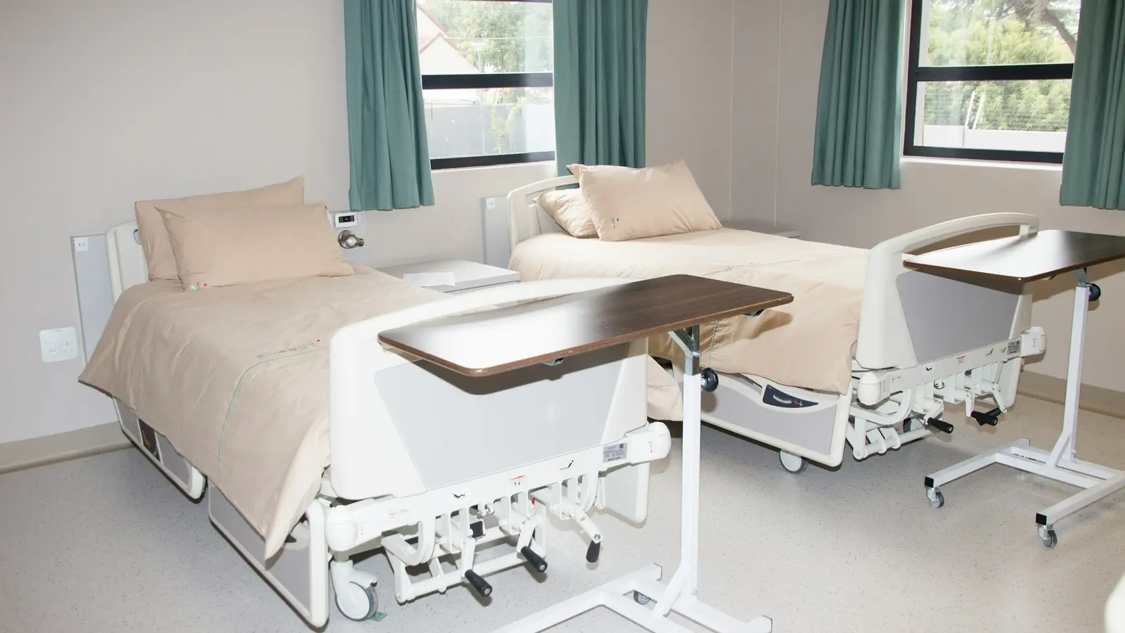 Made hospital beds with overbed tables beside teal-curtained windows