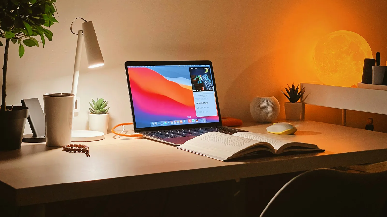 A home desk in warm evening light with an open laptop, an open notebook, a small white desk lamp and a glowing orange lamp beside a potted plant