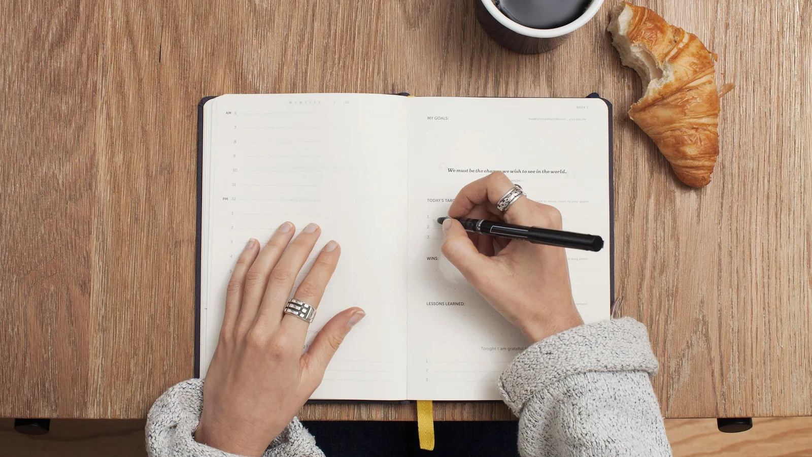 Two hands writing with a pen in an open ruled planner on a wooden table beside a cup of black coffee and a croissant, seen from above