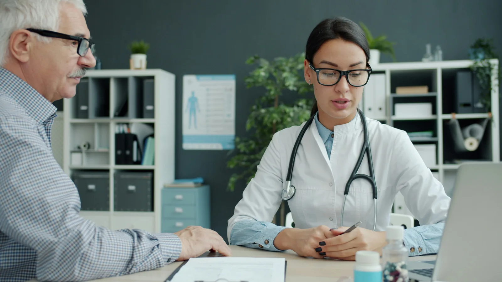 A general practitioner and a man in high-visibility workwear at a consulting room desk in natural light, the patient lifting his arm to shoulder height while the doctor watches, a teal folder and a blank form on the desk between them