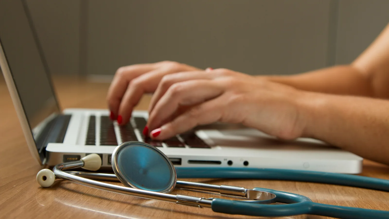 A clinician's hands typing on a laptop at a desk, a stethoscope coiled beside the keyboard in warm daylight