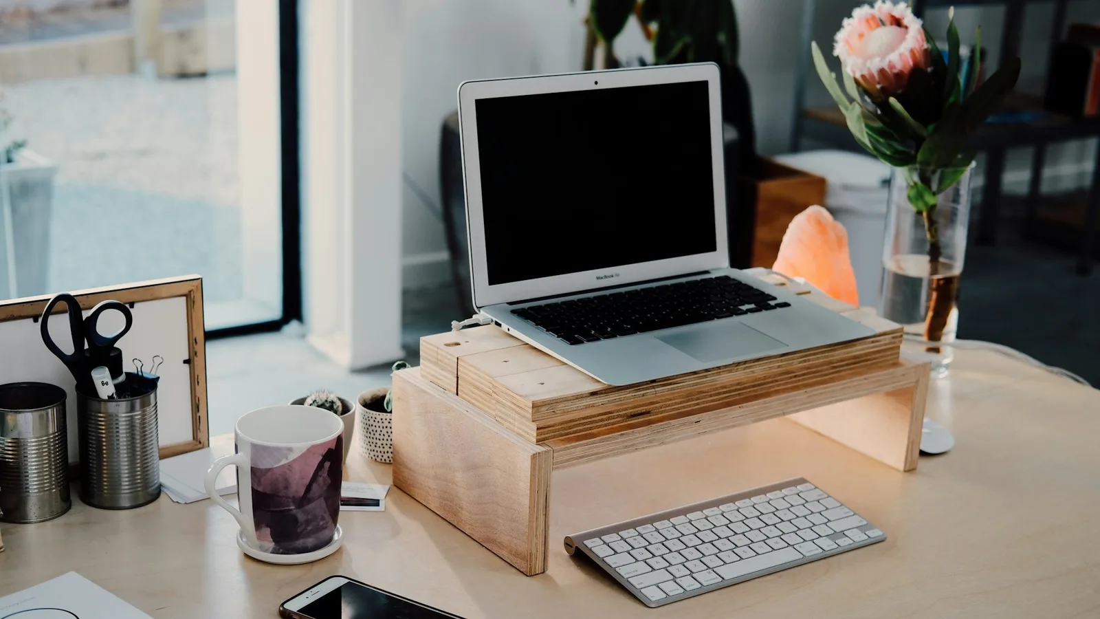 A tidy desk with a laptop on a timber stand, a wireless keyboard, a phone and a protea in a vase