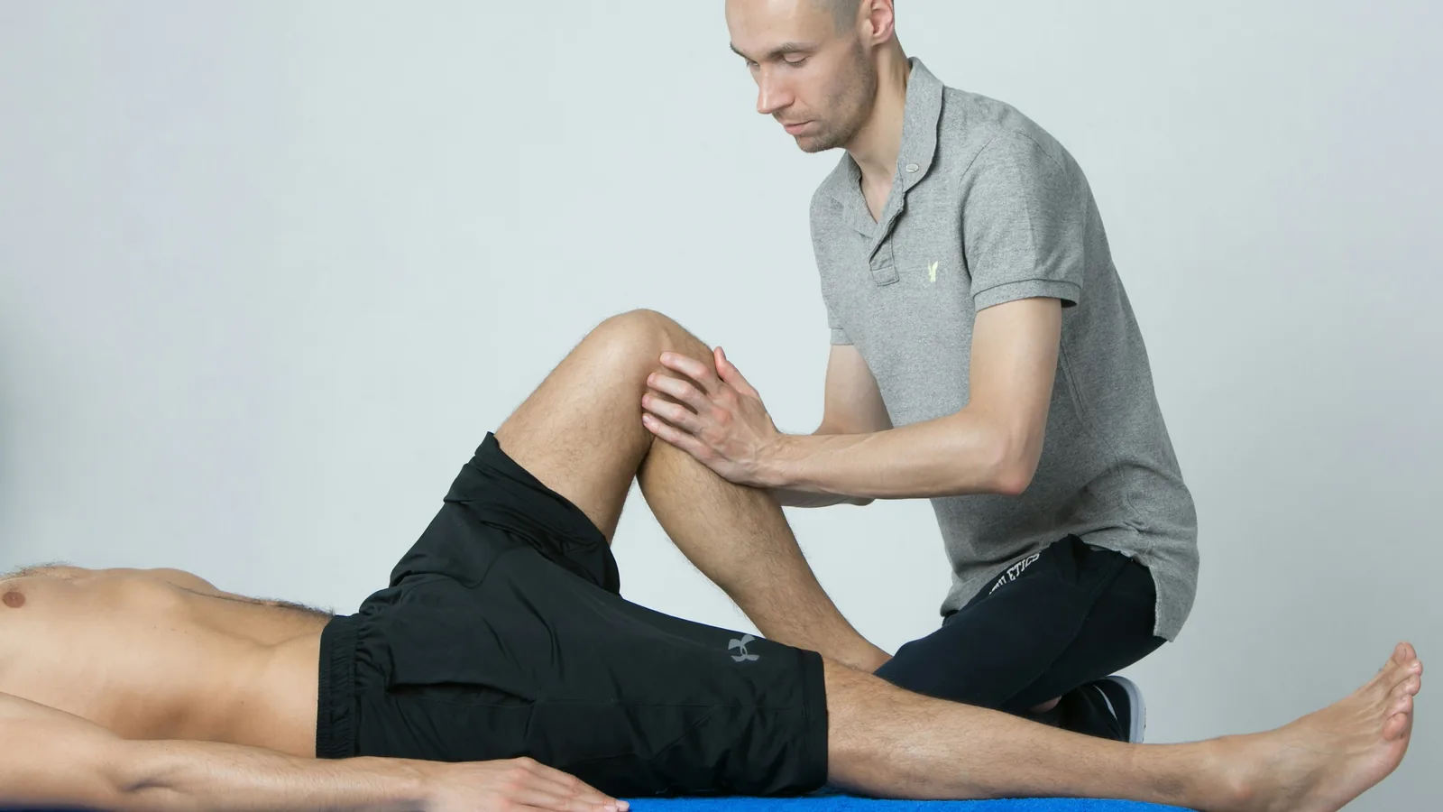 An Australian allied health treatment room in afternoon light, a physiotherapist's hands steadying a participant at the edge of a plinth partway through a transfer, a teal resistance band and an open notebook on the bench behind them