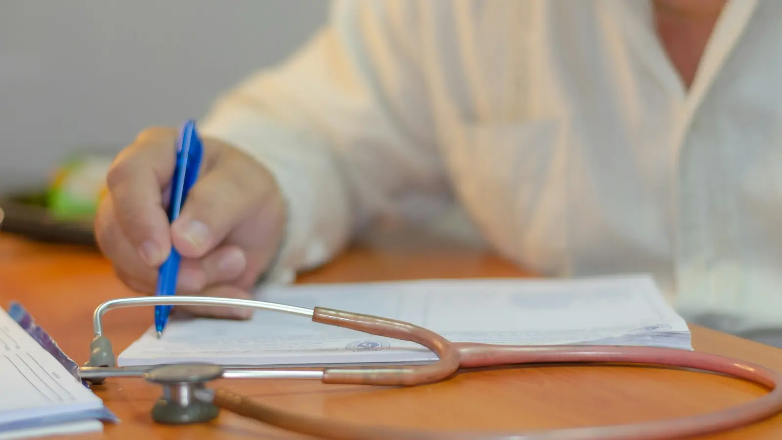 A general practitioner at a consulting room desk reads a printed letter beside a bright window