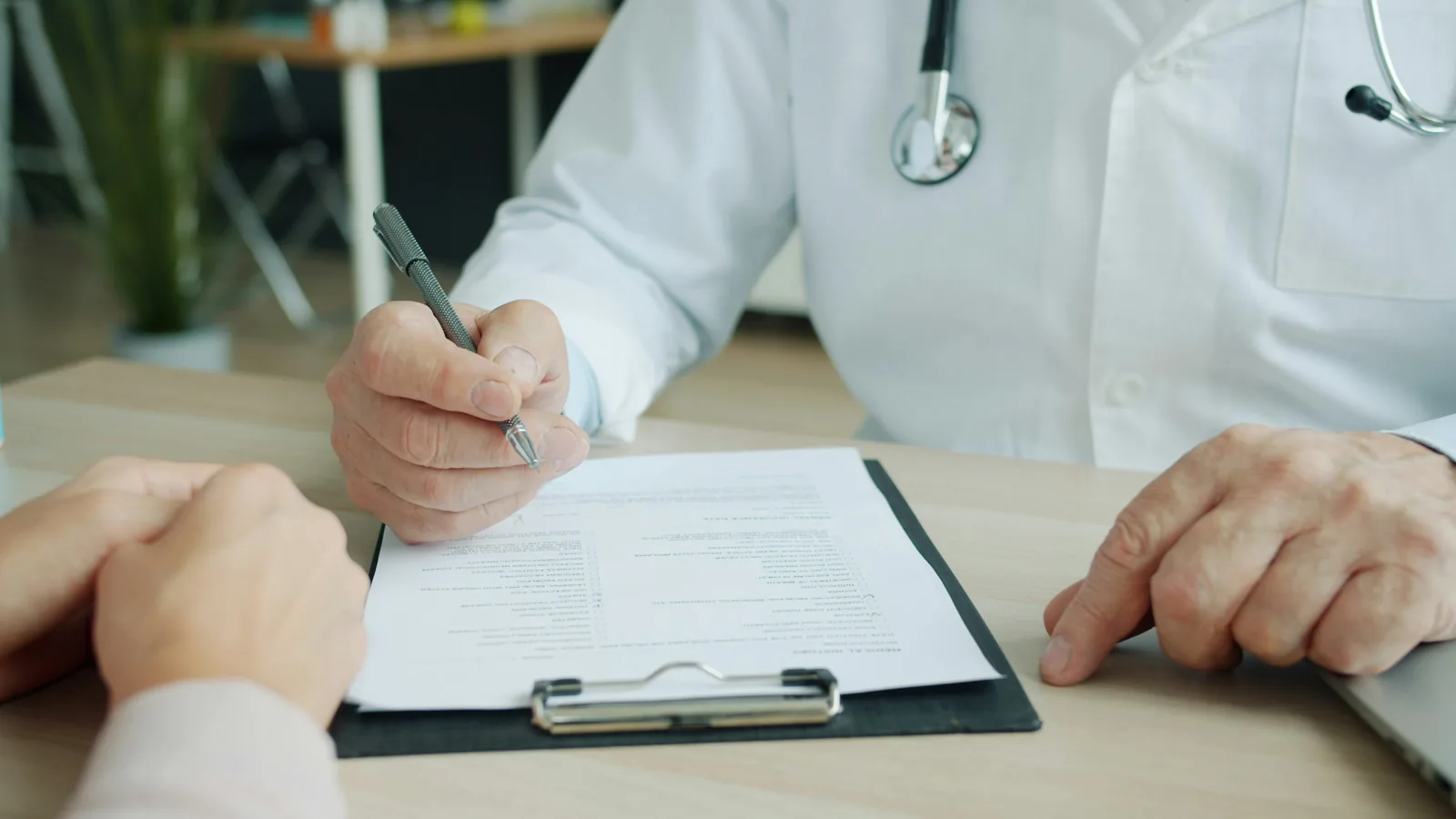 A clinician writing on a clipboard across the desk from a patient whose hands rest on the table
