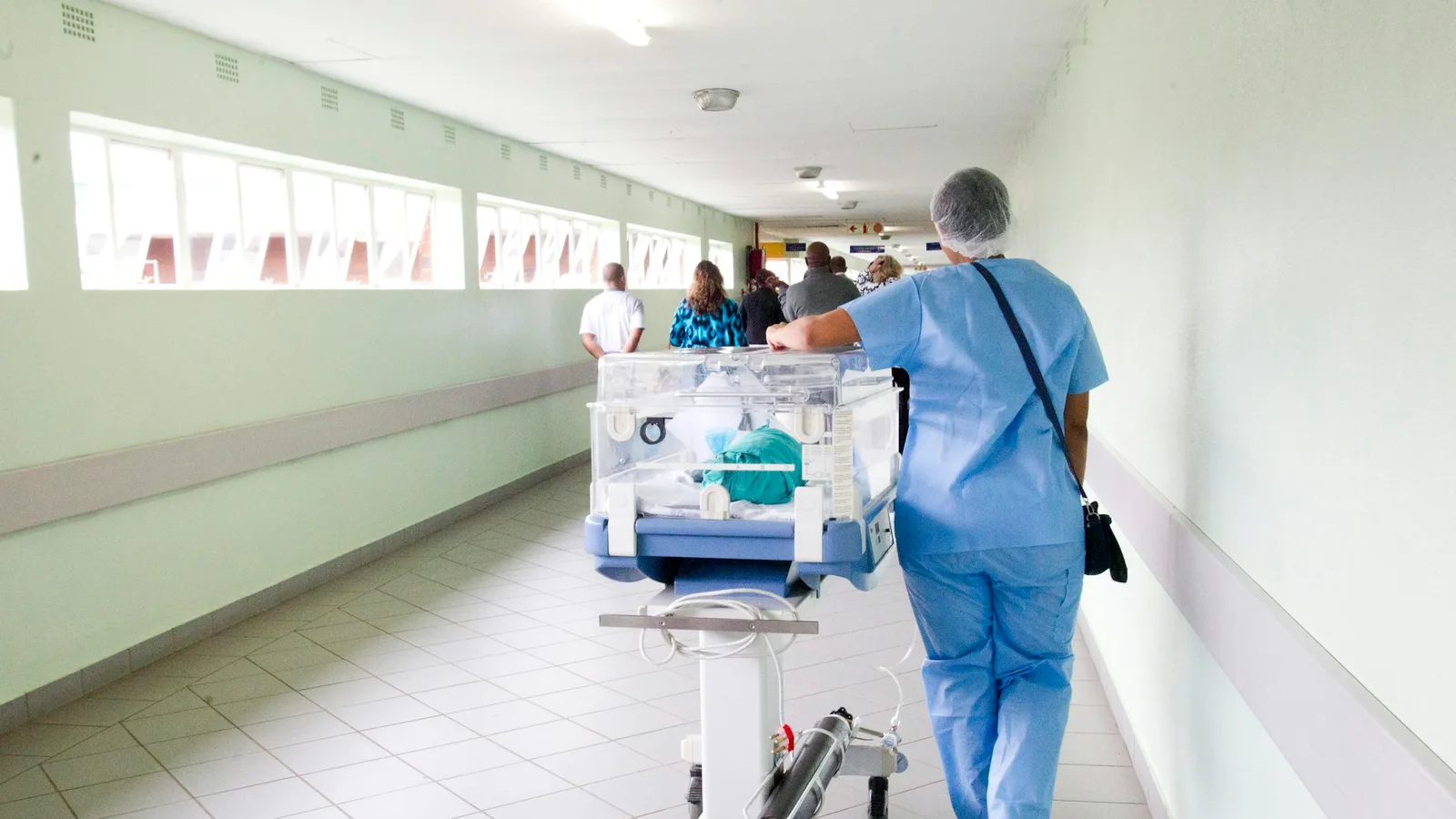 A nurse in scrubs pushes an infant incubator down a bright hospital corridor