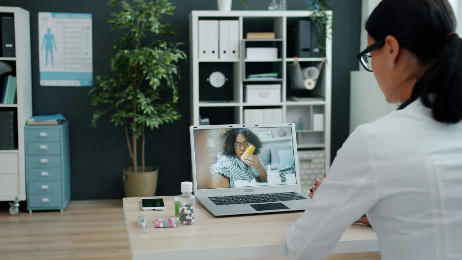 A clinician seen from behind takes a video call on a laptop at a sunlit desk, a stethoscope coiled beside the keyboard