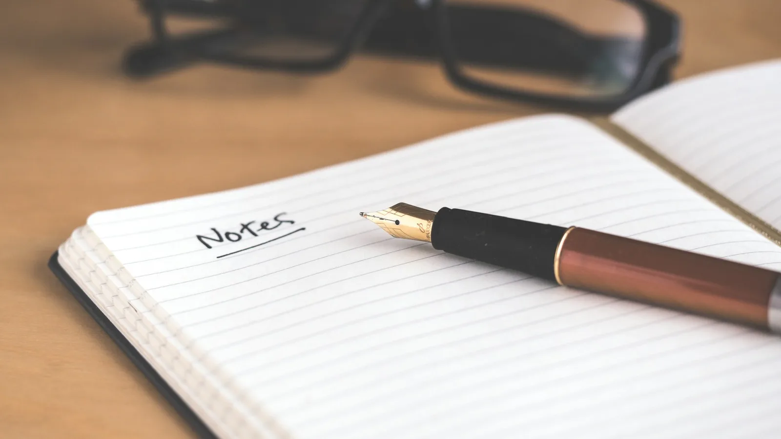 A fountain pen rests on an open lined notebook whose page is headed with the handwritten, underlined word 'Notes', with a pair of glasses soft-focused on the wooden desk behind