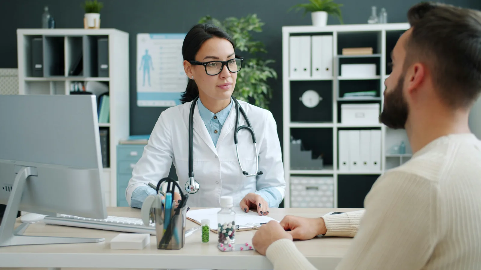 A clinician at a consulting room desk turned away from the monitor toward the patient sitting opposite